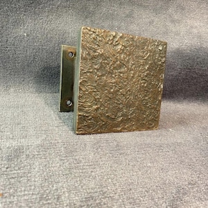 Könnte beinhalten: Vierkantiger Türgriff aus Bronze mit strukturierter Oberfläche. Der Griff hat eine rechteckige Montageplatte mit zwei Schraubenlöchern. Der quadratische Griff hat ein detailliertes, wirbelndes Muster.