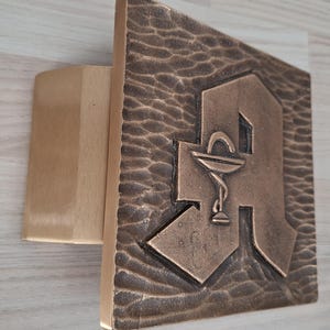 Peut inclure: Une plaque murale carrée de couleur bronze avec une surface texturée et martelée. La plaque présente une grande lettre A en relief avec un petit symbole de caducée embossé, représentant un emblème médical ou pharmaceutique, au centre.