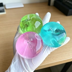 May include: Three translucent apple-shaped objects in green, pink, and teal are held in a gloved hand. Each apple has a stem and a smooth, glossy surface. The objects are likely decorative or artistic pieces.