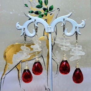 May include: Dangling earrings with clear quartz and teardrop-shaped red gemstones. The earrings are suspended from silver-colored hooks and are displayed on a white decorative stand. The background features a gold and white deer illustration.