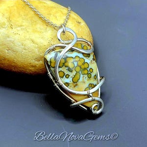 May include: A wire-wrapped pendant necklace featuring a triangular stone with a cream, light blue, and mustard yellow spotted pattern. The stone is encased in silver-toned metal wire coils and hangs from a matching silver-toned chain.