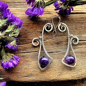 May include: A pair of handcrafted silver earrings with a wire-wrapped design. Each earring features a teardrop shape with a vibrant purple amethyst bead. The earrings are set against a rustic wooden background, with purple flowers.