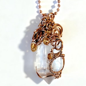 May include: A handcrafted pendant featuring a clear quartz crystal wrapped in copper wire. The wirework has intricate swirls and holds small, yellow-orange gemstones. The pendant hangs from a copper-colored ball chain, creating a unique, artistic accessory.