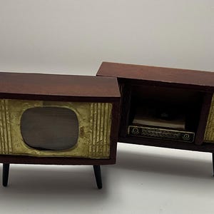 Puede incluir: Miniatura de televisión y radio de estilo vintage. Los armarios de madera son de color marrón oscuro con detalles dorados y patas negras. El televisor tiene una pantalla redondeada, mientras que la radio tiene un compartimento abierto con una pequeña radio en su interior.