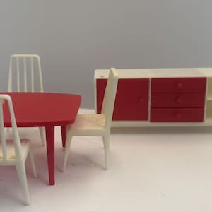 Puede incluir: Un juego de comedor en miniatura para casa de muñecas que incluye una mesa rectangular roja con cuatro sillas de color blanco hueso con listones verticales, y un aparador a juego con puertas y cajones rojos junto a estantes abiertos.
