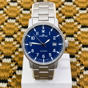 May include: A stainless steel Fortis automatic watch with a blue dial. The watch features white numerals, hands, and a date display at the 3 o'clock position. The watch is displayed on a white stand.