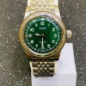 Puede incluir: Un reloj de pulsera de acero inoxidable con esfera verde. El reloj tiene una esfera redonda con números y agujas blancas, y una pantalla de fecha. La correa del reloj es de acero inoxidable y tiene un diseño texturizado. El reloj se exhibe sobre un soporte blanco.