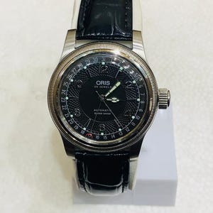 Può includere: Un orologio da polso con quadrante nero, cassa color argento e cinturino in pelle nera. Il quadrante dell'orologio presenta un motivo strutturato, lancette luminose e il testo "ORIS 25 JEWELS AUTOMATIC SWISS MADE". L'orologio è esposto su una superficie bianca.