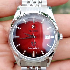 May include: A silver-toned Rado Golden Horse watch with a red dial. The watch features a date window at the 3 o'clock position and a silver-toned band. The dial has silver-toned hour markers and hands. The watch is Swiss made.