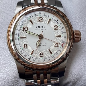 Può includere: Un orologio da polso automatico Oris con quadrante bianco, lunetta color oro rosa e bracciale a maglie in acciaio inossidabile bicolore argento e oro rosa. Il quadrante presenta un centro testurizzato, numeri arabi e la scritta Oris 25 Jewels.