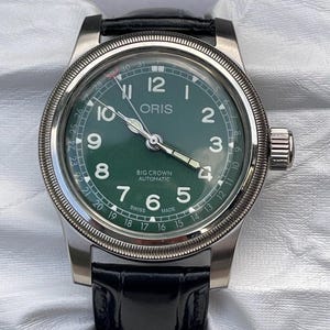 Puede incluir: Primer plano de un reloj Oris Big Crown Automatic con esfera verde y agujas plateadas. El reloj tiene una caja de acero inoxidable y una correa de cuero negro. La esfera muestra la hora y la fecha.