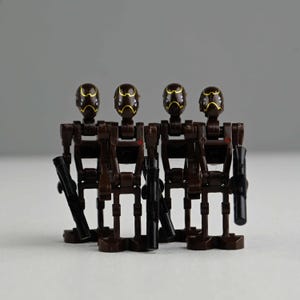 May include: Four brown and black toy robot figures with gold accents on their heads and black blasters. The figures are standing close together against a gray background. The robots have a distinct, angular design.