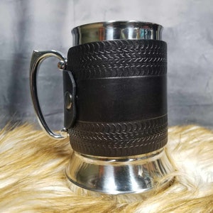 May include: A stainless steel beer mug with a black leather wrap. The leather has a textured pattern and a band around the middle.