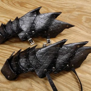 May include: A pair of black leather arm guards featuring a layered, scale-like texture. Each guard is constructed from overlapping pointed segments secured with silver-toned metal rivets and includes adjustable black leather straps with metal buckles.