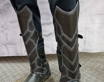 May include: A pair of black leather leg guards with a geometric pattern. The guards are worn over black pants and have buckles and straps.
