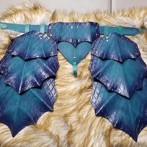 May include: A teal and blue leather dragon scale belt with gold accents. The belt has a front piece with a pointed shape and two side pieces with multiple layers of scales.