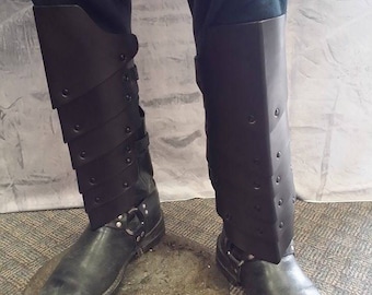 May include: Black leather leg armor with multiple straps and buckles. The armor is worn over black pants and boots.