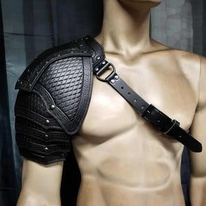 May include: Black leather shoulder armor with a diamond-patterned design and riveted accents. The armor includes a strap that goes across the chest. This piece is designed to protect the shoulder and upper arm.