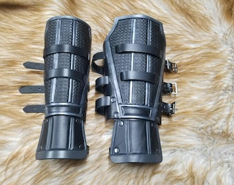 May include: A pair of black forearm guards featuring a textured basketweave pattern on the central panels, smooth black borders, and three adjustable leather straps with metal buckles on the side for secure fastening around the arm.
