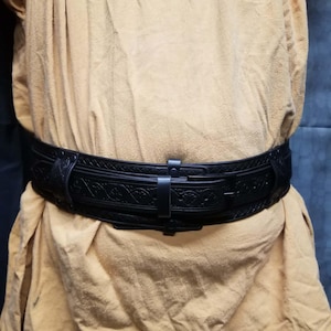 May include: A black leather belt featuring an intricate embossed floral pattern across the strap. The belt is secured with a matching black metal buckle and a single belt loop, worn over a textured, mustard-yellow fabric garment.