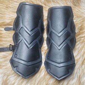 May include: A pair of black forearm guards featuring a segmented, layered design with raised ridges and pointed edges. Each guard includes two adjustable black straps with metal buckles on the side for secure fastening around the arm.