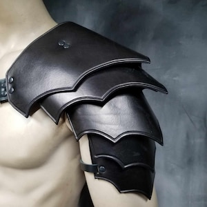 Op de afbeelding: Zwarte leren schouderbescherming met een gelaagd ontwerp. De bescherming heeft een band die om de borst gaat en een band die om de biceps gaat.