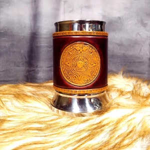 May include: A stainless steel mug with a brown leather wrap featuring a gold Aztec calendar design.
