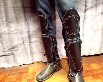 May include: A pair of black leather greaves and knee guards featuring a segmented design with visible rivets and adjustable straps. The protective armor covers the lower legs and knees, worn over dark trousers and paired with dark leather boots.