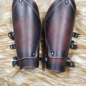 May include: A pair of dark brown leather arm guards with decorative edging and rivets. Each guard has three black buckles and straps for securing. The leather has a gradient effect, transitioning from dark brown to a reddish hue.