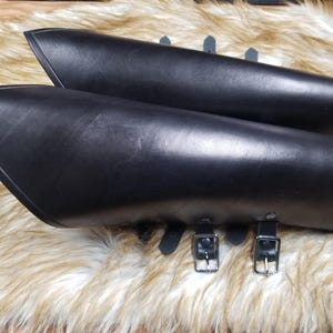 May include: A pair of black leather arm guards with adjustable straps and buckles. The guards are made of thick leather and have a smooth finish.