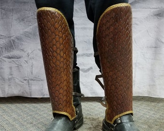 May include: A pair of brown leather leg guards with gold trim. The guards are made of a textured leather and have a strap at the top.