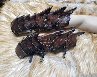 May include: A pair of brown leather gauntlets featuring a textured scale pattern, layered plates, and brass-colored metal rivets. The gauntlets are secured with black leather straps and small brass-colored metal buckles on the underside.