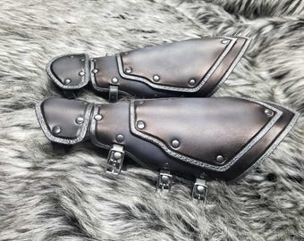 May include: A pair of dark gray forearm guards featuring a layered design with metallic silver rivets and textured edges. Each guard is secured with three adjustable silver-toned metal buckles and straps, providing a structured fit for the lower arm.