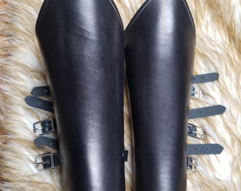 May include: A pair of black leather bracers featuring a pointed top edge and three adjustable straps with silver-toned metal buckles on the side. The smooth, dark material is shaped to fit the forearm with a curved bottom edge.