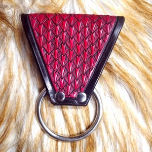 May include: A red leather ring holder with a scale pattern and a silver ring. The holder is triangular in shape and has two silver rivets.