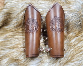 May include: A pair of brown leather arm guards with a tree of life design. The guards are secured with straps and buckles.