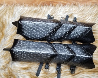 May include: A pair of black leather arm guards with a scale-like pattern. The guards are secured with straps and buckles.