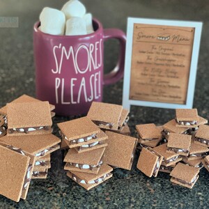 May include: A pile of miniature s'mores, each made with two graham crackers, a layer of chocolate and marshmallow. The s'mores are arranged on a brown surface, with a mug that says "S'mores Please!" and a sign that says "S'more Menu" in the background.