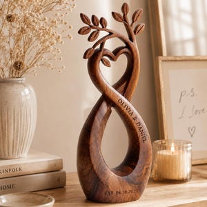 May include: A dark brown wooden sculpture featuring an infinity loop design that forms a heart shape at the top with carved leaves on the branches. The wood is engraved with the text OLIVIA & DANIEL and the date EST. 06.18.2022 on the base.