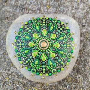 Peut inclure: Un galet de rivière clair et lisse orné d'un motif mandala peint à la main. L'œuvre se compose de cercles concentriques complexes, de formes en goutte d'eau et de points dans des tons vifs de vert citron, vert forêt et or sur fond noir.