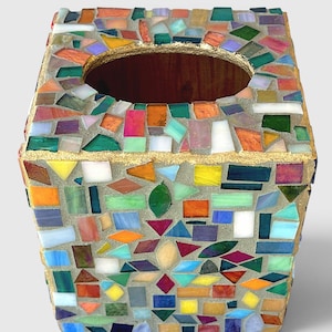 May include: A square tissue box cover featuring a multi-coloured mosaic design made of small, irregularly shaped glass pieces in shades of red, orange, yellow, green, blue, and purple, set in grey grout with a gold-painted top edge and opening.