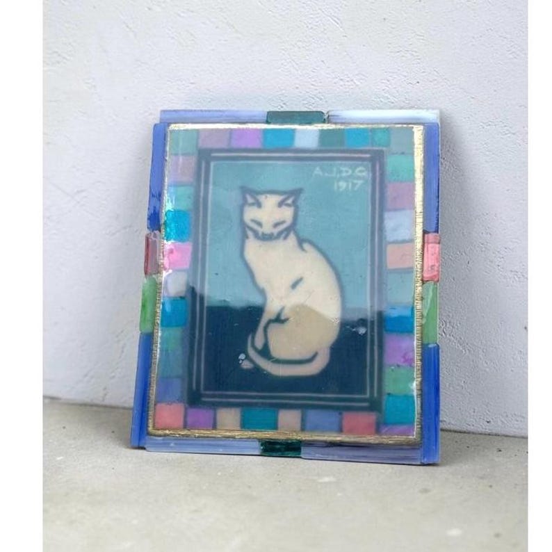 May include: A rectangular glass art piece featuring a central illustration of a seated cream-colored cat against a teal and dark blue background. The image includes the text A.J.D.G. 1917 and is framed by a border of small, multicolored glass tiles.