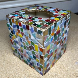K&ouml;nnte beinhalten: Ein quadratischer Taschentuchbox-&Uuml;berzug, verziert mit einem bunten Mosaik aus kleinen Glasfliesen in Rot, Blau, Gr&uuml;n, Gelb und Wei&szlig;. Die Kanten und die ovale &Ouml;ffnung an der Oberseite sind mit einem goldfarbenen Rand auf wei&szlig;em Fugenm&ouml;rtel versehen.