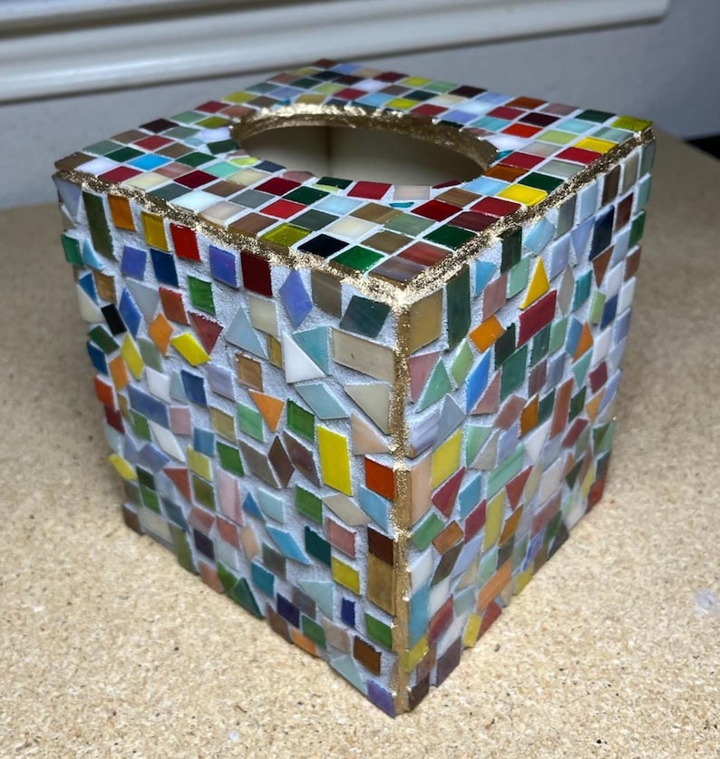 K&ouml;nnte beinhalten: Ein quadratischer Taschentuchbox-&Uuml;berzug, verziert mit einem bunten Mosaik aus kleinen Glasfliesen in Rot, Blau, Gr&uuml;n, Gelb und Wei&szlig;. Die Kanten und die ovale &Ouml;ffnung an der Oberseite sind mit einem goldfarbenen Rand auf wei&szlig;em Fugenm&ouml;rtel versehen.