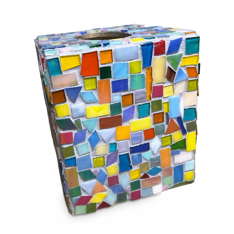 May include: A square-shaped tissue box cover decorated with a mosaic of small, multi-colored glass tiles in shades of red, yellow, blue, green, orange, and white, all set into a light gray grout base with a circular opening at the top.