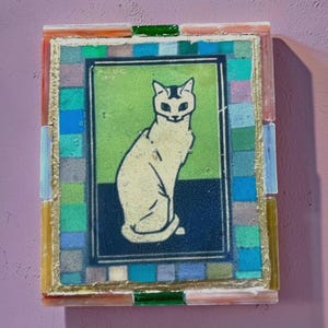 May include: A rectangular mosaic wall art piece featuring a central illustration of a seated cat in cream against a green and dark blue background. The artwork is framed by a border of small, multicolored glass tiles in blue, green, and grey.
