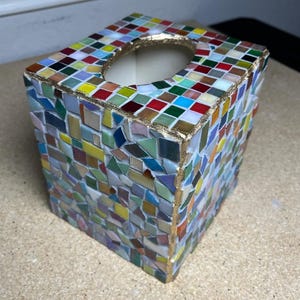 K&ouml;nnte beinhalten: Ein quadratischer Taschentuchbox-&Uuml;berzug mit einem mehrfarbigen Mosaikfliesenmuster. Die Au&szlig;enseite ist mit kleinen, unregelm&auml;&szlig;ig geformten Glasfliesen in Rot-, Blau-, Gr&uuml;n-, Gelb- und Wei&szlig;t&ouml;nen verziert, mit goldfarbenen Kanten am oberen Rand.