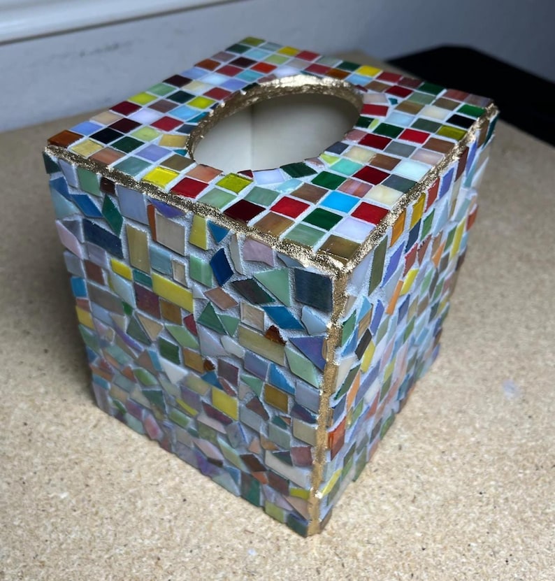 K&ouml;nnte beinhalten: Ein quadratischer Taschentuchbox-&Uuml;berzug mit einem mehrfarbigen Mosaikfliesenmuster. Die Au&szlig;enseite ist mit kleinen, unregelm&auml;&szlig;ig geformten Glasfliesen in Rot-, Blau-, Gr&uuml;n-, Gelb- und Wei&szlig;t&ouml;nen verziert, mit goldfarbenen Kanten am oberen Rand.