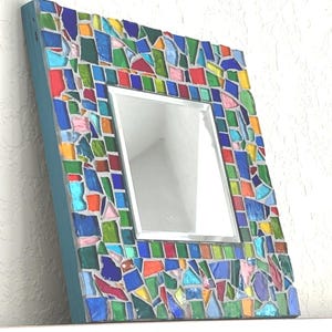 May include: A square wall mirror featuring a frame decorated with a mosaic of multi-colored glass pieces in shades of blue, green, red, yellow, and pink. The glass fragments are set in gray grout, and the side of the frame is painted teal blue.