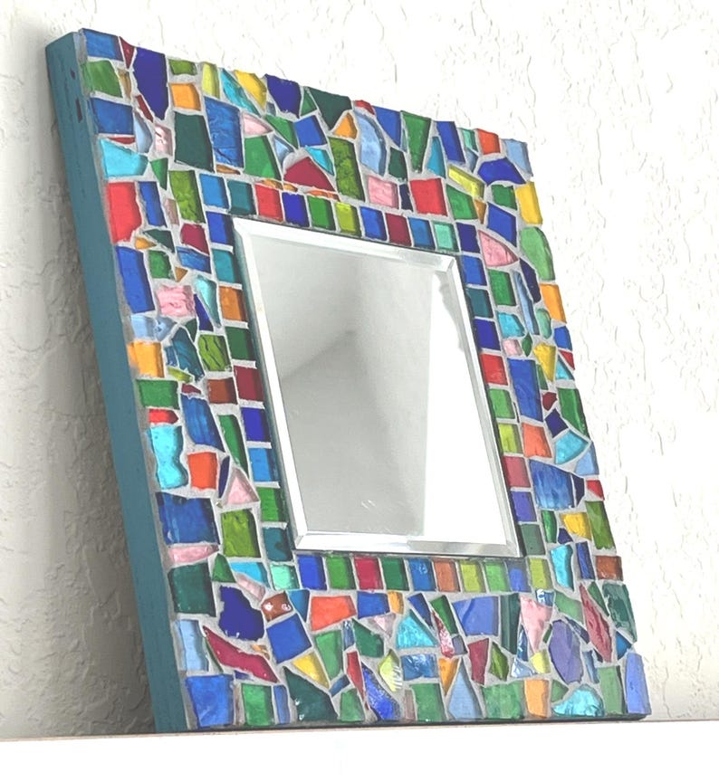 May include: A square wall mirror featuring a frame decorated with a mosaic of multi-colored glass pieces in shades of blue, green, red, yellow, and pink. The glass fragments are set in gray grout, and the side of the frame is painted teal blue.